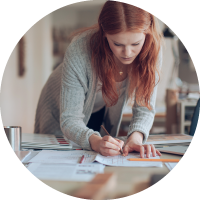 A woman leans over her cluttered desk and writes on a sheet of paper. 